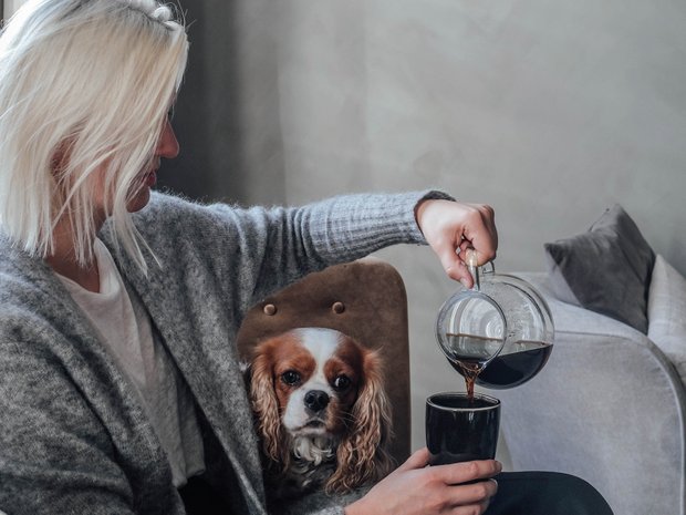 Woman sits on a couch with a dog and pours coffee into a cup.